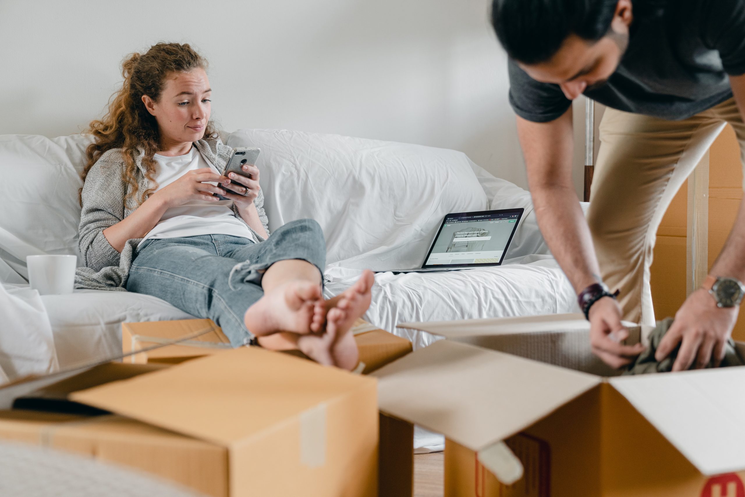 picture of woman sitting on couch next to laptop using iphone and man standing unpacking a moving box