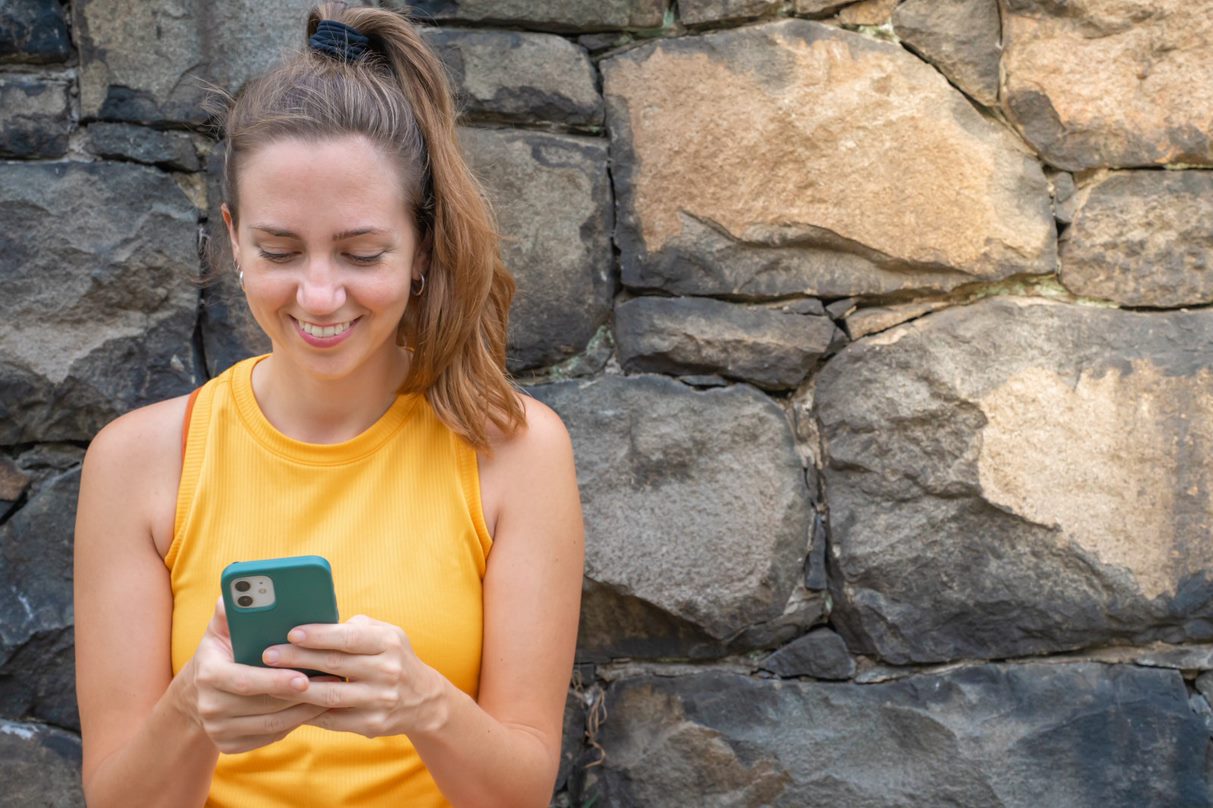A young person looking at their phone while leaning against a wall of rocks learning about how to use conversational marketing social media in multifamily with the ever-growing popularity of social media with prospective renters