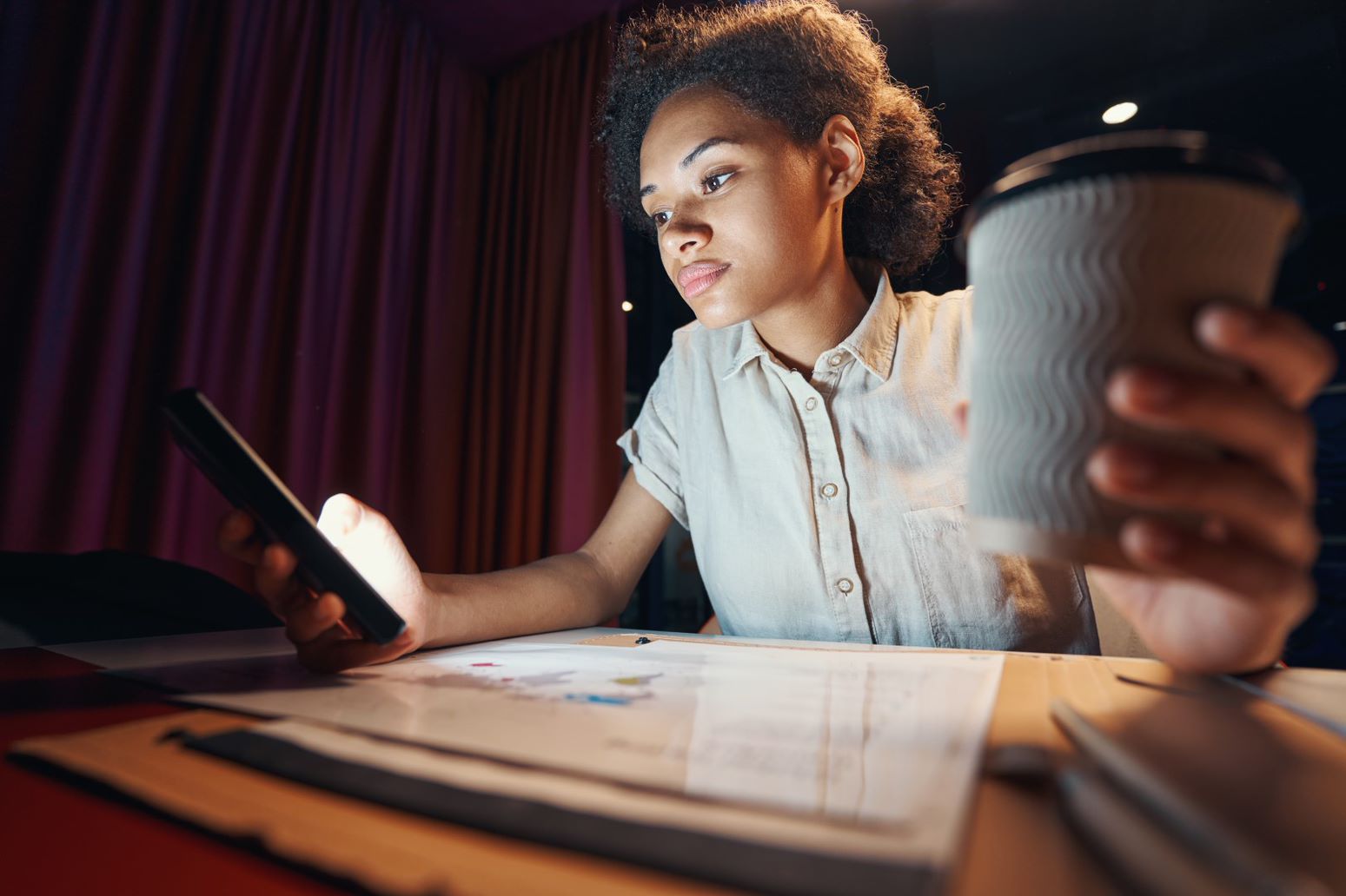A young person sitting at a desk while looking at her phone and holding a cup of coffee while reading about how to prepare for the upcoming multifamily market outlook
