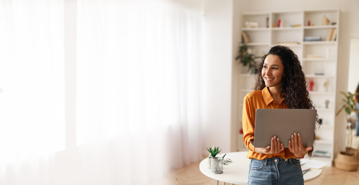 Smiling woman holding an open laptop and looking out a window