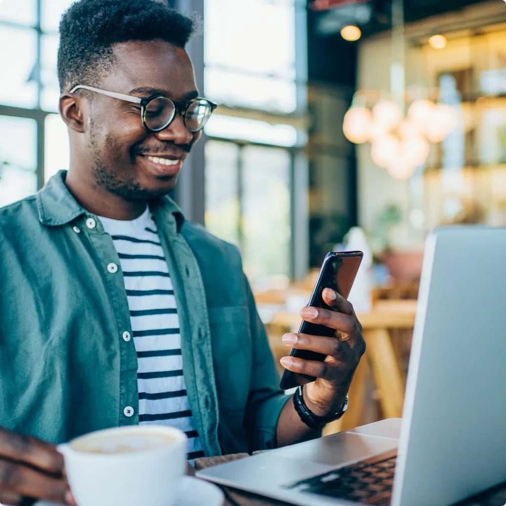 Multifamily Websites | PERQ AI Leasing Assistant A man smiles while looking at his phone, with a coffee on the other hand. He's facing a laptop.