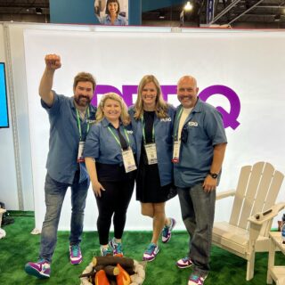PERQ booth at the Apartmentalize multifamily conference, featuring four team members in matching blue shirts posing in front of branded signage, a cozy setup with white Adirondack chairs, and a faux campfire displayed on green carpeting.