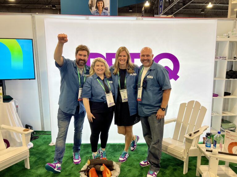 PERQ booth at the Apartmentalize multifamily conference, featuring four team members in matching blue shirts posing in front of branded signage, a cozy setup with white Adirondack chairs, and a faux campfire displayed on green carpeting.