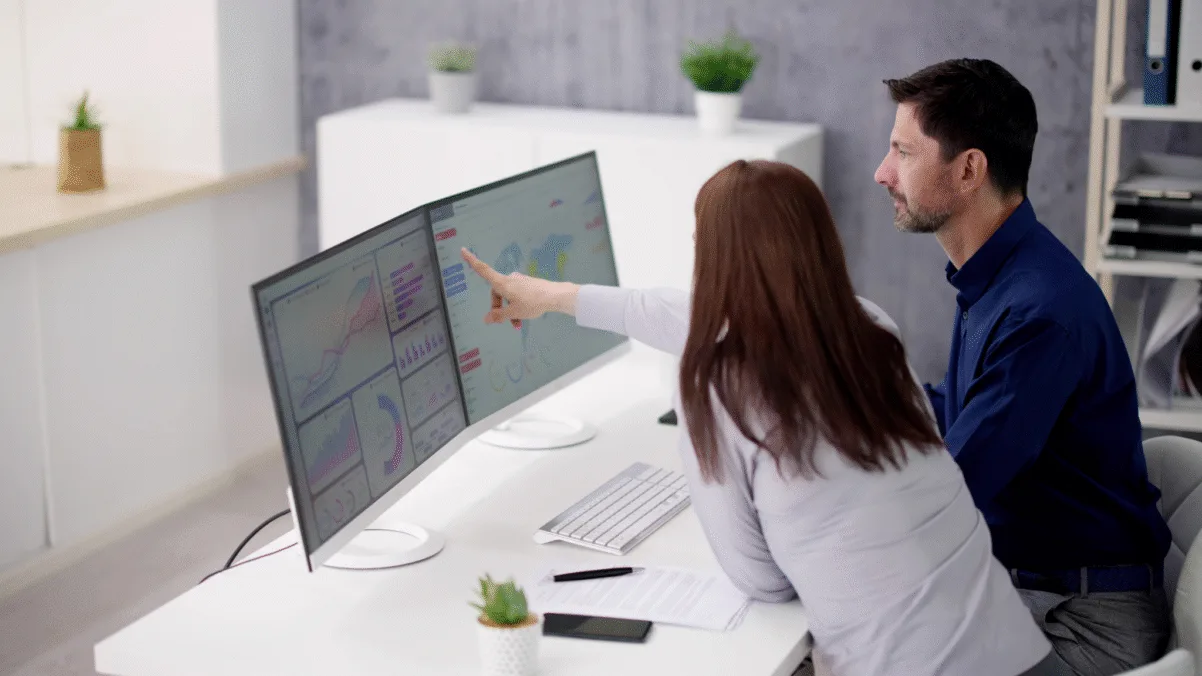 Two people sitting at a desk and looking at a two-screen desktop together.