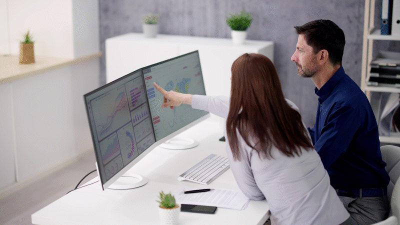 Two people sitting at a desk and looking at a two-screen desktop together.